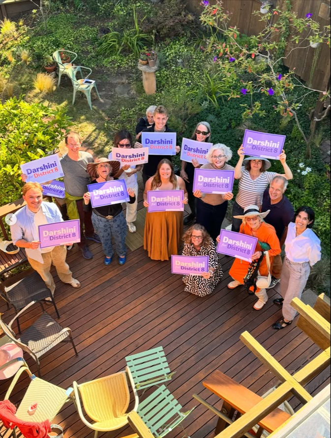 Neighbors attending Wendy's Garden Party for District 8 Supervisor Candidate Darshini Patel gather and hold campaign signs, smiling in front of a garden backdrop.